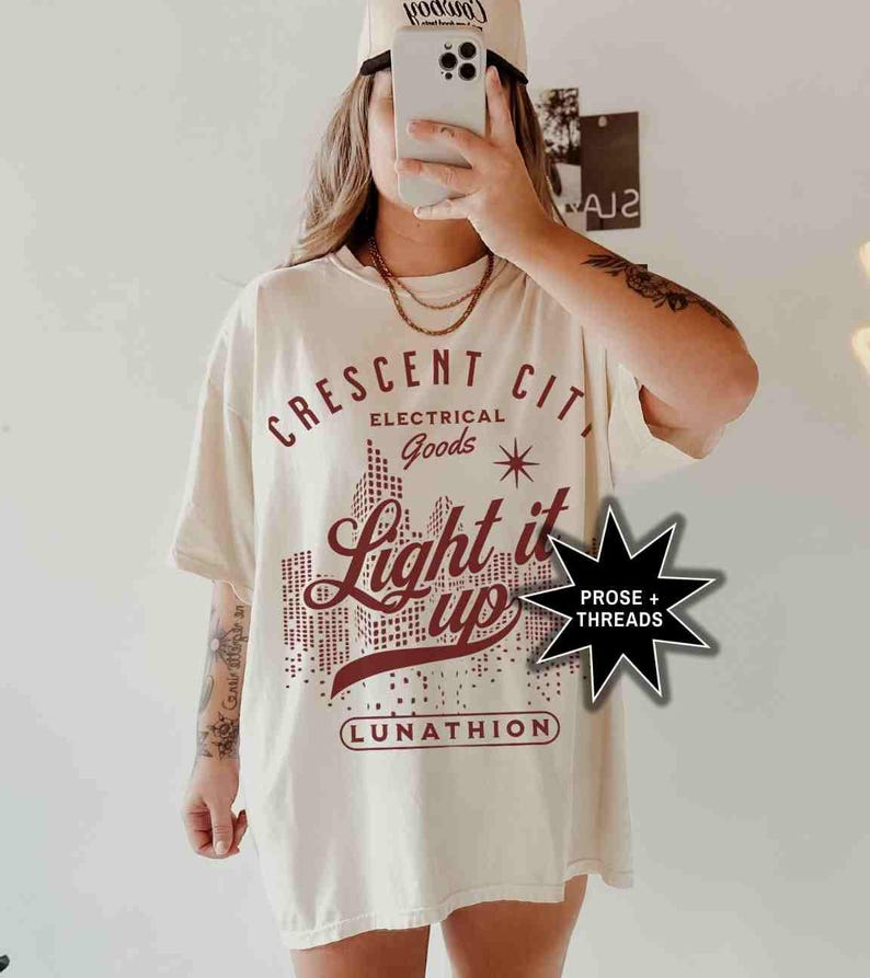 May include: A white t-shirt with a red graphic design that says "Crescent City Electrical Goods Light it Up" with a star and a city skyline. The graphic also includes the text "Lunathion" and "Prose + Threads".