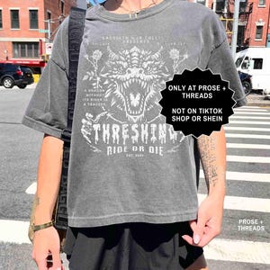 May include: Gray graphic t-shirt featuring a white dragon design with the text "THRESHING RIDE OR DIE EST. 2023". The shirt also includes the text "A DRAGON WITHOUT ITS RIDER IS A TRAGEDY".