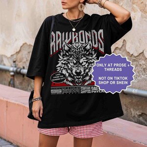 Op de afbeelding: Zwart grafisch T-shirt met een wolf ontwerp en de tekst "RAWHORDS". Het shirt heeft ook de tekst "ONLY AT PROSE + THREADS NOT ON TIKTOK SHOP OR SHEIN". Het model draagt rood-witte geruite shorts.