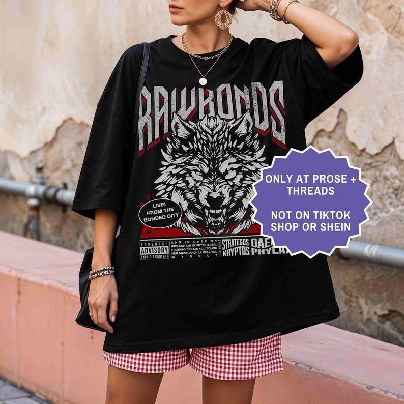 Op de afbeelding: Zwart grafisch T-shirt met een wolf ontwerp en de tekst "RAWHORDS". Het shirt heeft ook de tekst "ONLY AT PROSE + THREADS NOT ON TIKTOK SHOP OR SHEIN". Het model draagt rood-witte geruite shorts.