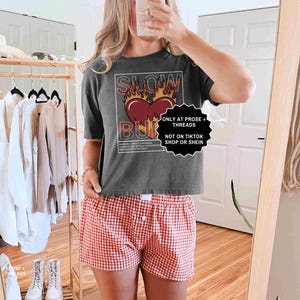 May include: A dark gray t-shirt with the text "SLOW BURN" over a heart design with flames. The person is wearing red and white checkered shorts. A clothing rack with various garments is in the background.