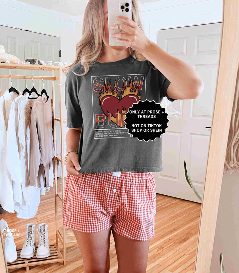 May include: A dark gray t-shirt with the text "SLOW BURN" over a heart design with flames. The person is wearing red and white checkered shorts. A clothing rack with various garments is in the background.