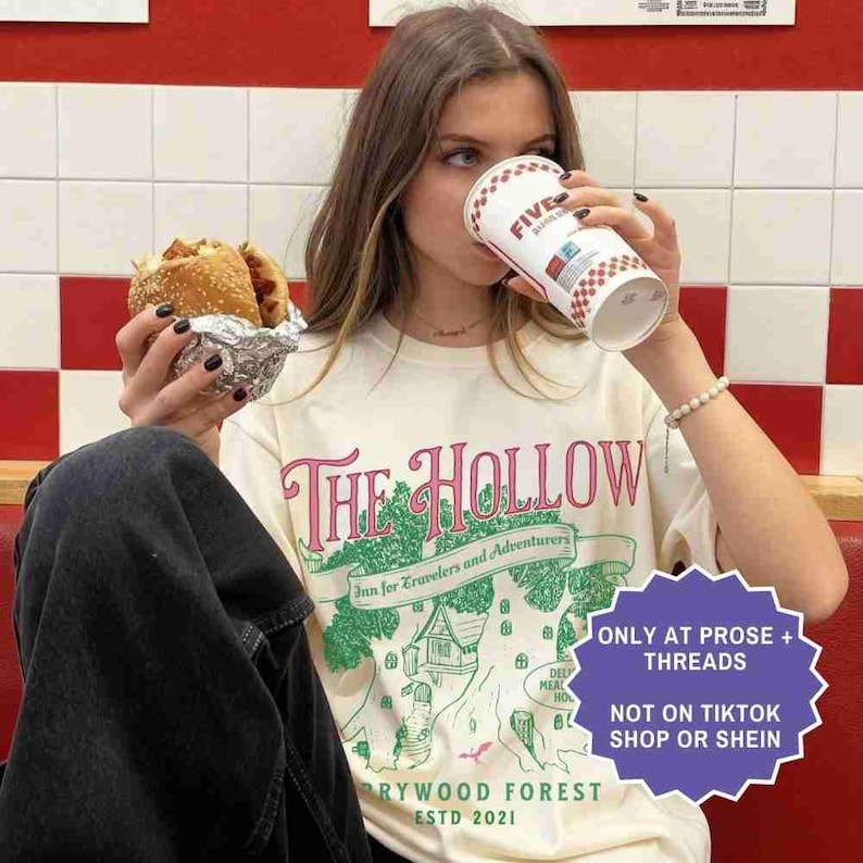 Pu&ograve; includere: T-shirt color crema con una grafica che recita "The Hollow Inn for Travelers and Adventurers" in rosa e verde. L'immagine include anche un hamburger avvolto nella carta stagnola e un bicchiere Five Guys. Un adesivo viola dice "ONLY AT PROSE + THREADS" e "NOT ON TIKTOK SHOP OR SHEIN".