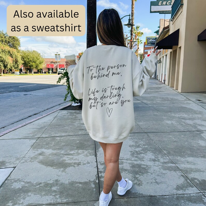 May include: Cream-colored sweatshirt with black text on the back that reads: "To the person behind me, Life is tough my darling, but so are you." A small heart is drawn below the text. The text "Also available as a sweatshirt" is in the upper left corner.