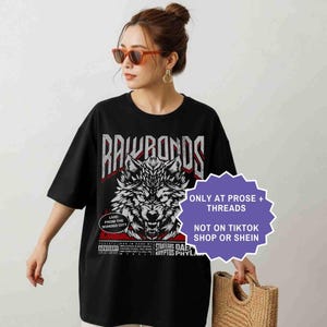 Op de afbeelding: Zwart T-shirt met een wolfafbeelding en de tekst "RAWRANDS". Het shirt heeft ook de tekst "LIVE! FROM THE BONDED CITY" en "ONLY AT PROSE + THREADS NOT ON TIKTOK SHOP OR SHEIN".
