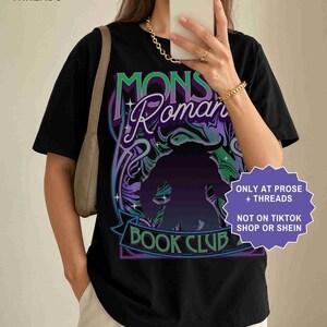 Op de afbeelding: Zwart t-shirt met een grafisch ontwerp met de tekst "MONS Roman BOOK CLUB" in paars en groen. Het ontwerp bevat een silhouet van een monster. Het shirt heeft ook de tekst "ONLY AT PROSE + THREADS" en "NOT ON TIKTOK SHOP OR SHEIN".