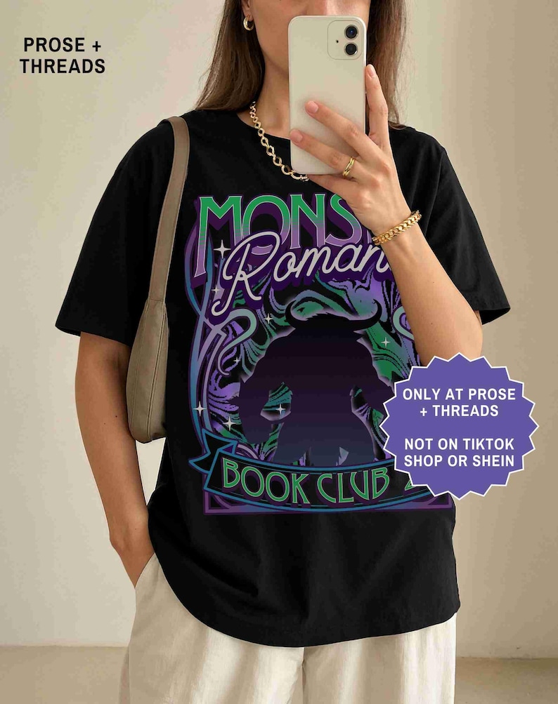 Op de afbeelding: Zwart t-shirt met een grafisch ontwerp met de tekst "MONS Roman BOOK CLUB" in paars en groen. Het ontwerp bevat een silhouet van een monster. Het shirt heeft ook de tekst "ONLY AT PROSE + THREADS" en "NOT ON TIKTOK SHOP OR SHEIN".