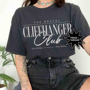 May include: Black t-shirt with white text that reads "The Brutal Cliffhanger Club" with the words "Still Crying" and "Still Waiting" below. A black star with white text that reads "Prose + Threads" is in the upper right corner.