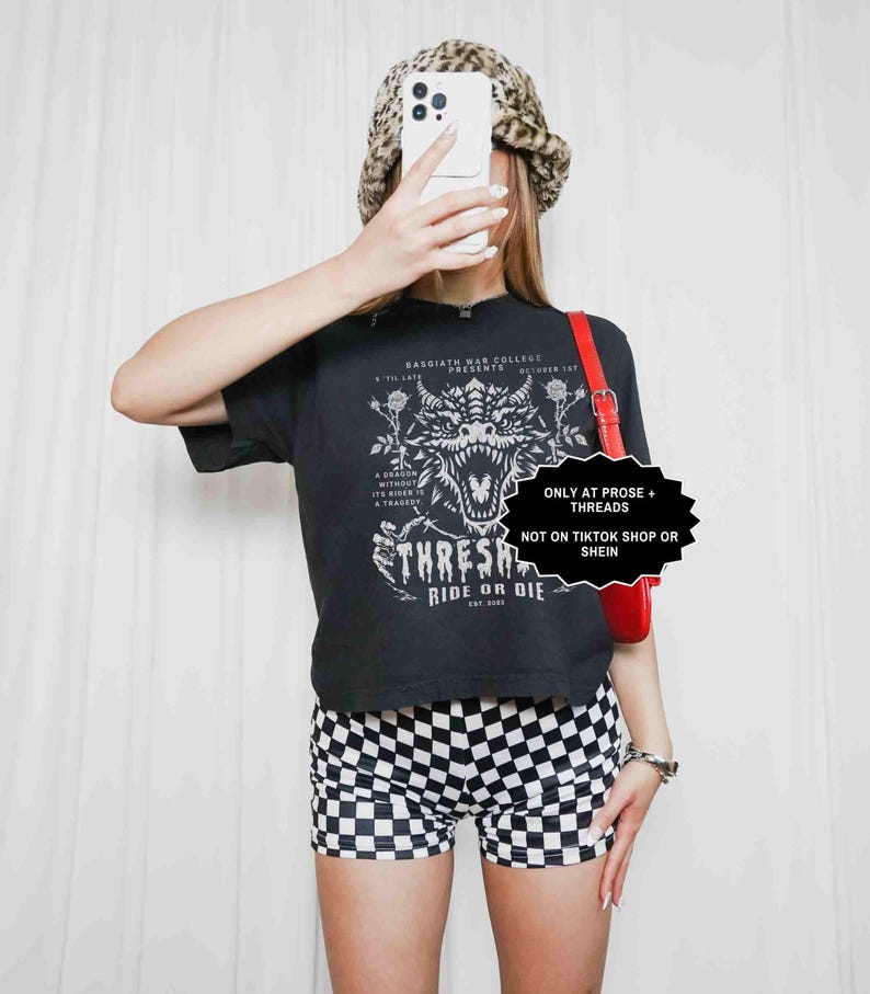 May include: A black t-shirt with a dragon graphic and the text "THRESH RIDE OR DIE". The model is wearing black and white checkered shorts, a leopard print hat, and a red purse. The image also contains the text "ONLY AT PROSE + THREADS NOT ON TIKTOK SHOP OR SHEIN".