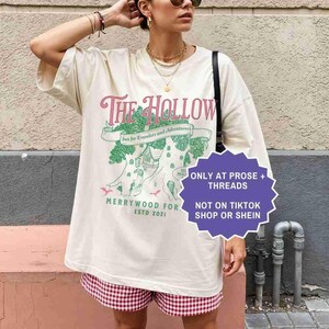 Pu&ograve; includere: T-shirt oversize color crema con la scritta "The Hollow" in rosa e verde, con una grafica di una casa sull'albero. La maglietta include anche il testo "Merrywood For Estd 2021". La modella indossa pantaloncini a quadri rossi e bianchi.