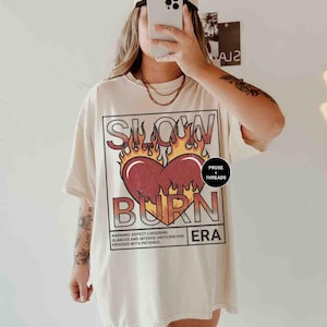 May include: A cream-colored oversized t-shirt with a graphic print of a red heart with flames and the text "Slow Burn Era". The graphic also includes the text "Prose + Threads".