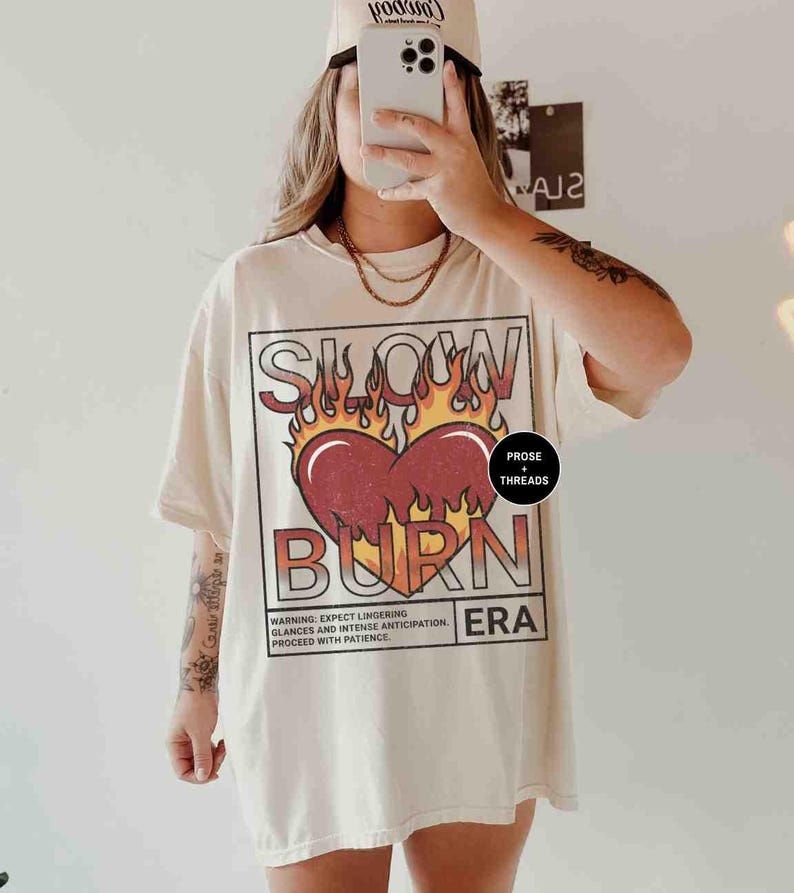 May include: A cream-colored oversized t-shirt with a graphic print of a red heart with flames and the text "Slow Burn Era". The graphic also includes the text "Prose + Threads".