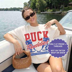 May include: Cream-colored t-shirt with the words "PUCK Buddies" in red and blue, featuring hockey player graphics. The shirt also has the text "HOCKEY ROMANCE CLUB". A brown woven handbag is held in the hand.
