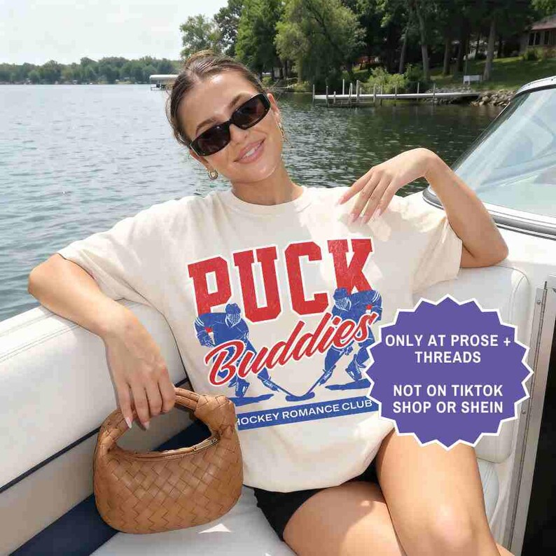 May include: Cream-colored t-shirt with the words "PUCK Buddies" in red and blue, featuring hockey player graphics. The shirt also has the text "HOCKEY ROMANCE CLUB". A brown woven handbag is held in the hand.