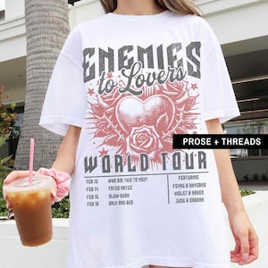 Pode incluir: Camiseta branca oversized com uma estampa gr&aacute;fica de um cora&ccedil;&atilde;o com rosas e o texto "Enemies to Lovers World Tour". O texto "Who Did This To You?" e uma lista de datas e atos musicais s&atilde;o impressos na parte inferior da camiseta.