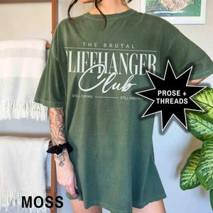May include: A green oversized t-shirt with the text "The Brutal Lifehanger Club" in white. The text "Still Crying" and "Still Waiting" are printed below the main text. The shirt has a distressed look and is made of a soft, comfortable fabric.