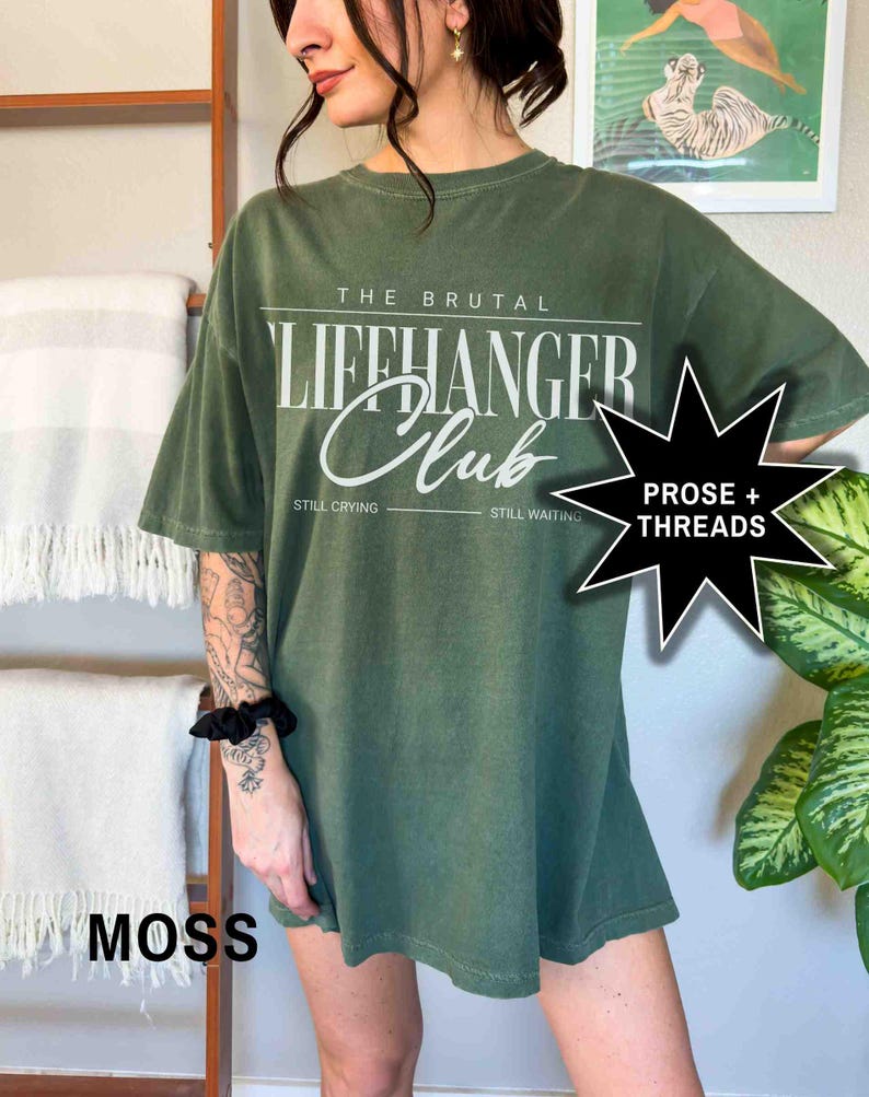 May include: A green oversized t-shirt with the text "The Brutal Lifehanger Club" in white. The text "Still Crying" and "Still Waiting" are printed below the main text. The shirt has a distressed look and is made of a soft, comfortable fabric.