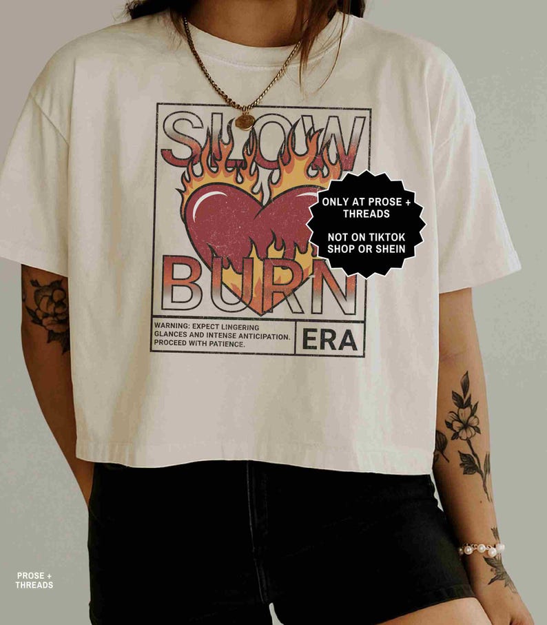 May include: White cropped t-shirt featuring a graphic design with the words "SLOW BURN" above a heart engulfed in flames. The shirt also includes the text "ERA" and a warning about lingering glances.