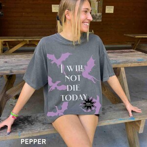 May include: A woman wearing a gray t-shirt with a purple dragon graphic and the text "I will not die today" and a black star with the text "Prose Threads".