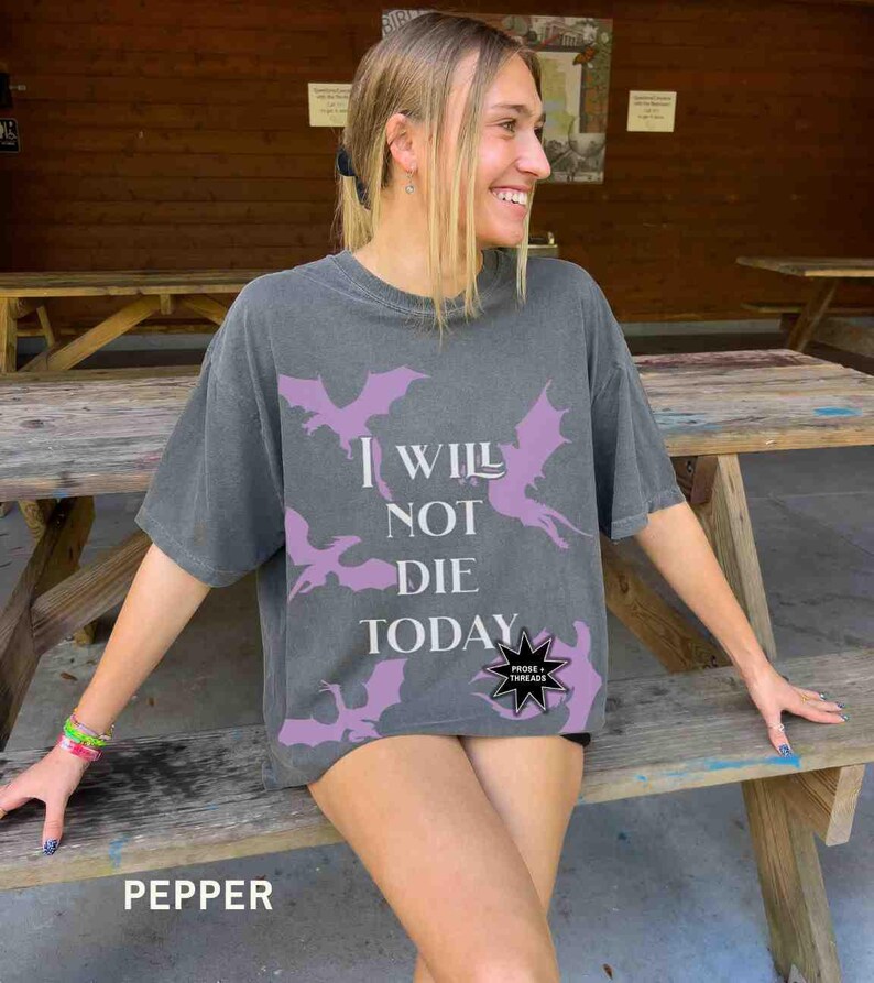May include: A woman wearing a gray t-shirt with a purple dragon graphic and the text "I will not die today" and a black star with the text "Prose Threads".
