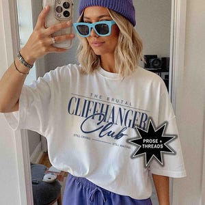 May include: White t-shirt with the text "The Brutal Cliffhanger Club" printed on it. The text is in a blue and black font. The shirt also has the text "Still Crying" and "Still Waiting" printed on it. The shirt is being worn by a person with a blue beanie and blue sunglasses. The image also includes a black star with the text "Prose + Threads" printed on it.