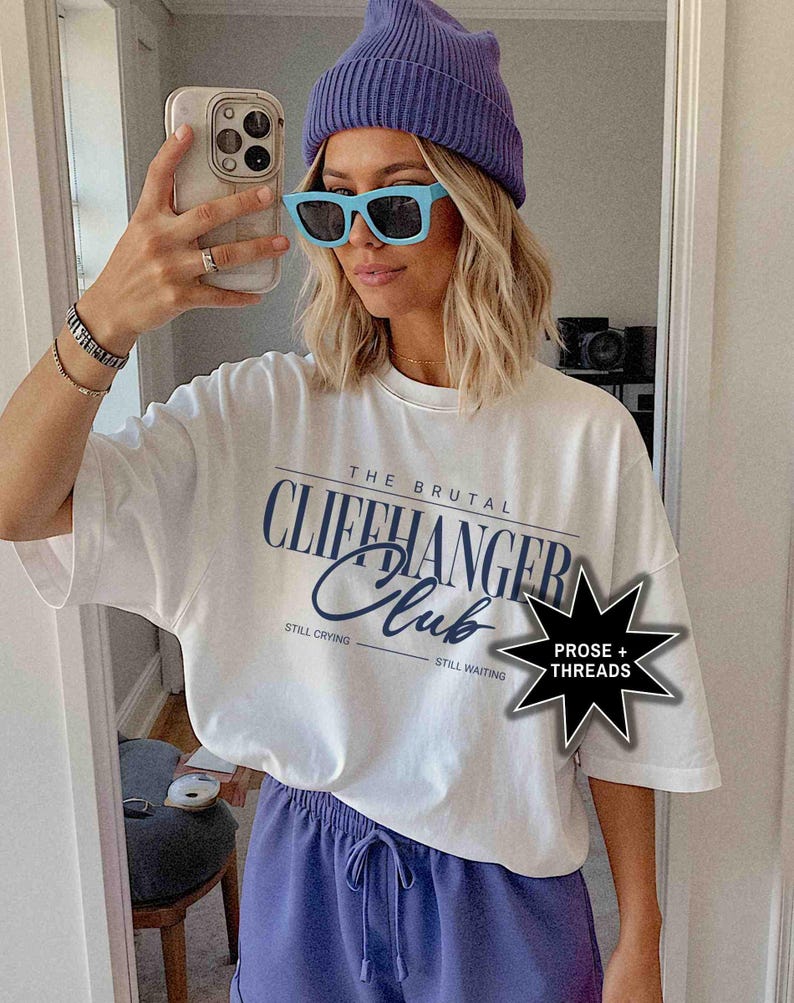 May include: White t-shirt with the text "The Brutal Cliffhanger Club" printed on it. The text is in a blue and black font. The shirt also has the text "Still Crying" and "Still Waiting" printed on it. The shirt is being worn by a person with a blue beanie and blue sunglasses. The image also includes a black star with the text "Prose + Threads" printed on it.