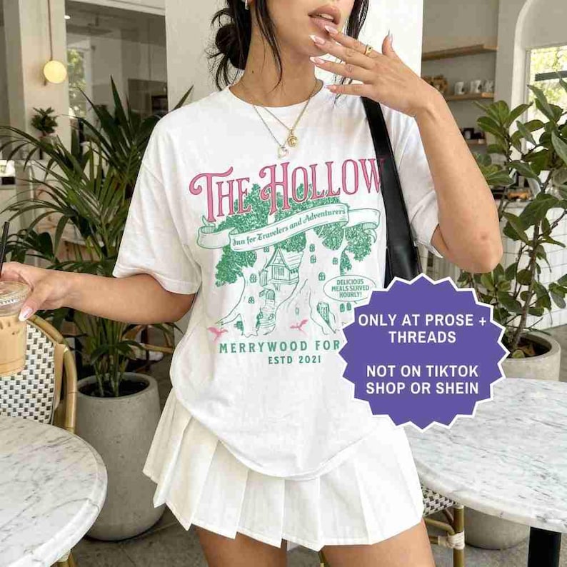 Pu&ograve; includere: T-shirt bianco con grafica rosa e verde con la scritta "The Hollow", con un'illustrazione di un albero e una casa. La maglietta ha anche il testo "Merrywood For Estd 2021". &Egrave; visibile anche una gonna a pieghe bianca.