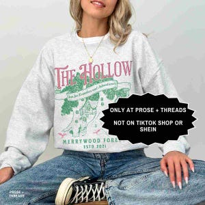 May include: A light grey sweatshirt with a pink and green graphic design. The design includes the text "The Hollow", a tree and house illustration, and the text "Inn for Travelers and Adventurers". The sweatshirt also has the text "MERRYWOOD FORE ESTD 2021".