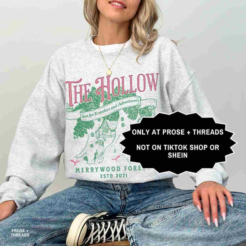 May include: A light grey sweatshirt with a pink and green graphic design. The design includes the text "The Hollow", a tree and house illustration, and the text "Inn for Travelers and Adventurers". The sweatshirt also has the text "MERRYWOOD FORE ESTD 2021".