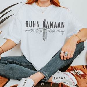 May include: White t-shirt with a black graphic print of a sword and the text "RUHN DANAAN knew three things with absolute certainty".