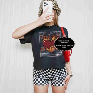 May include: Black t-shirt with a graphic design featuring a heart, flames, and the words "SLOW BURN ERA." The image also shows black and white checkered shorts and a leopard print bucket hat. A red purse is visible.