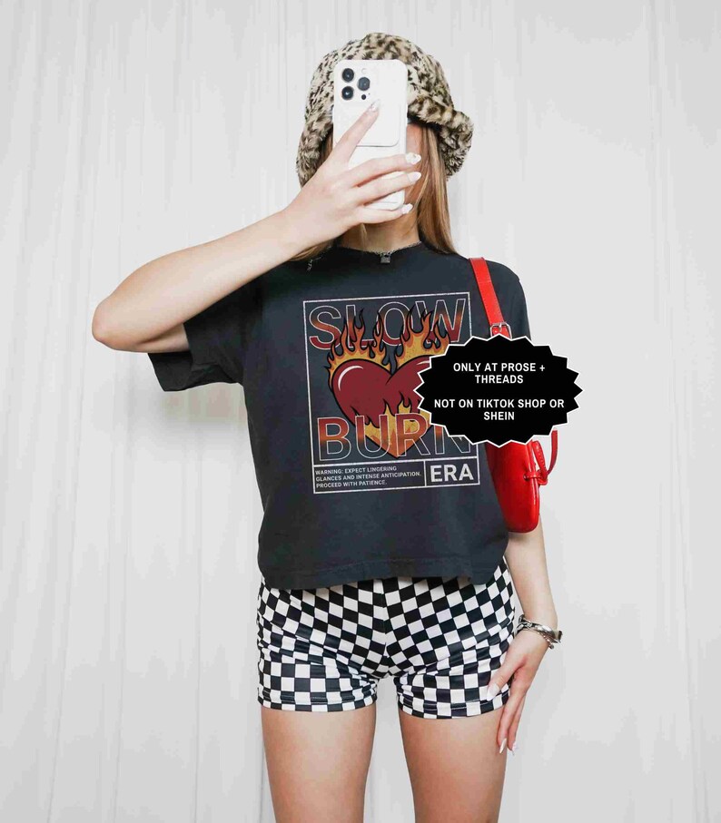 May include: Black t-shirt with a graphic design featuring a heart, flames, and the words "SLOW BURN ERA." The image also shows black and white checkered shorts and a leopard print bucket hat. A red purse is visible.