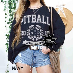 May include: A black sweatshirt with a white graphic print that says "Pitball" and "Zodiac Academy". The graphic also includes a wreath with the words "Solaris Champs" and the numbers "20" and "19". The sweatshirt is being worn by a person wearing denim shorts and a white belt.