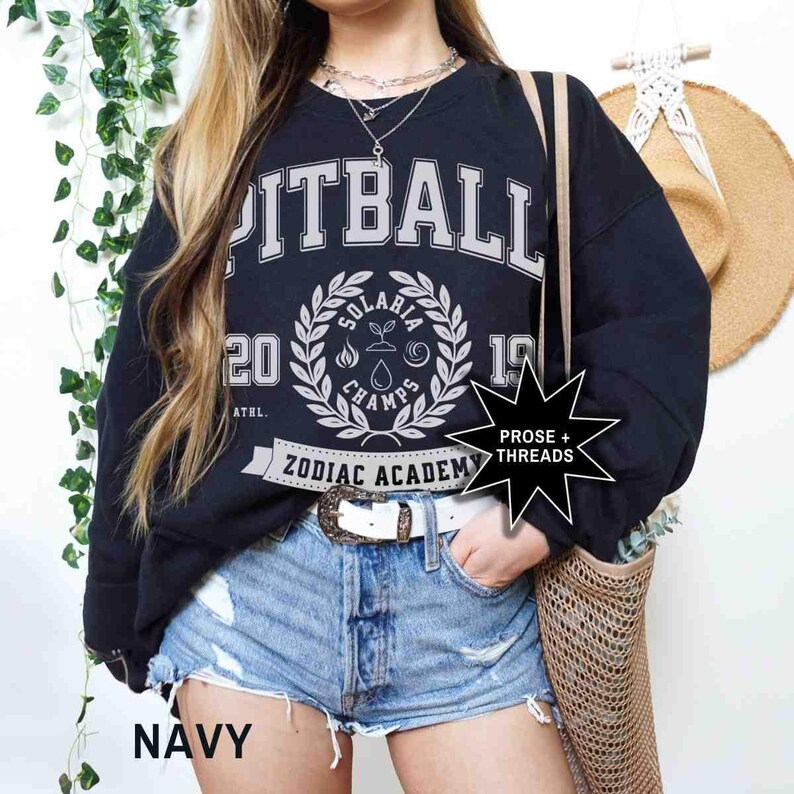 May include: A black sweatshirt with a white graphic print that says "Pitball" and "Zodiac Academy". The graphic also includes a wreath with the words "Solaris Champs" and the numbers "20" and "19". The sweatshirt is being worn by a person wearing denim shorts and a white belt.