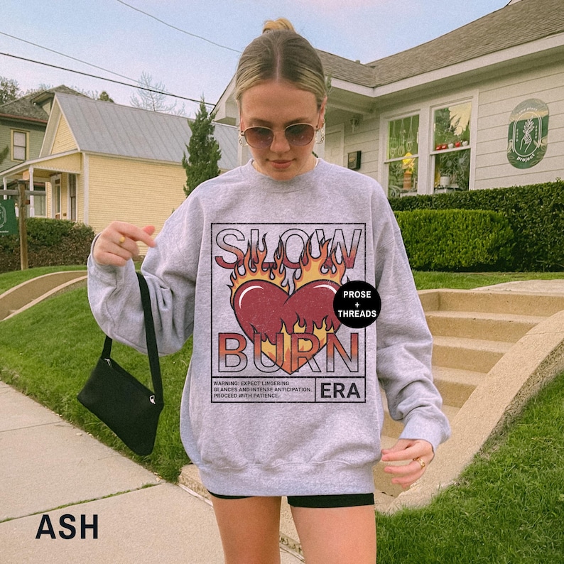 May include: A gray sweatshirt with a graphic print of a heart on fire with the text "Slow Burn" and "Prose + Threads" in a black rectangle. The text "Warning: Expect lingering glances and intense anticipation. Proceed with patience." is printed below the graphic. The text "ERA" is printed in a black rectangle below the warning text.