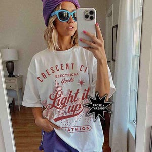 May include: White t-shirt with a red graphic that says "Crescent City Electrical Goods Light it Up" with a star and a city skyline. The graphic is in a pixelated style. The t-shirt also has a small black starburst sticker that says "Prose + Threads".