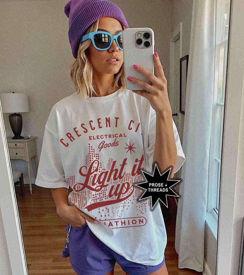 May include: White t-shirt with a red graphic that says "Crescent City Electrical Goods Light it Up" with a star and a city skyline. The graphic is in a pixelated style. The t-shirt also has a small black starburst sticker that says "Prose + Threads".