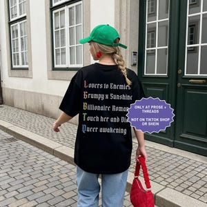 Pu&ograve; includere: Una t-shirt nera con un elenco verticale di tropi letterari in carattere gotico bianco, le cui prime lettere formano LGBTQ in colori arcobaleno. Il testo recita: Lovers to Enemies, Grumpy x Sunshine, Billionaire Romance, Touch her and Die, Queer awakening.