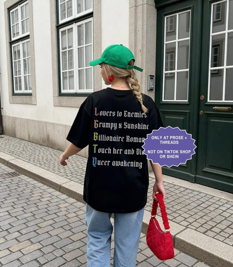 Pu&ograve; includere: Una t-shirt nera con un elenco verticale di tropi letterari in carattere gotico bianco, le cui prime lettere formano LGBTQ in colori arcobaleno. Il testo recita: Lovers to Enemies, Grumpy x Sunshine, Billionaire Romance, Touch her and Die, Queer awakening.