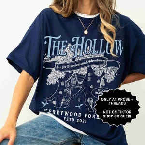 Pu&ograve; includere: T-shirt blu navy con colletto girocollo bianco. La maglietta presenta una grafica azzurra con la scritta "THE HOLLOW Inn for Travelers and Adventurers" e "MERRYWOOD FOR ESTD 2021". Un adesivo in bianco e nero &egrave; sul lato destro.