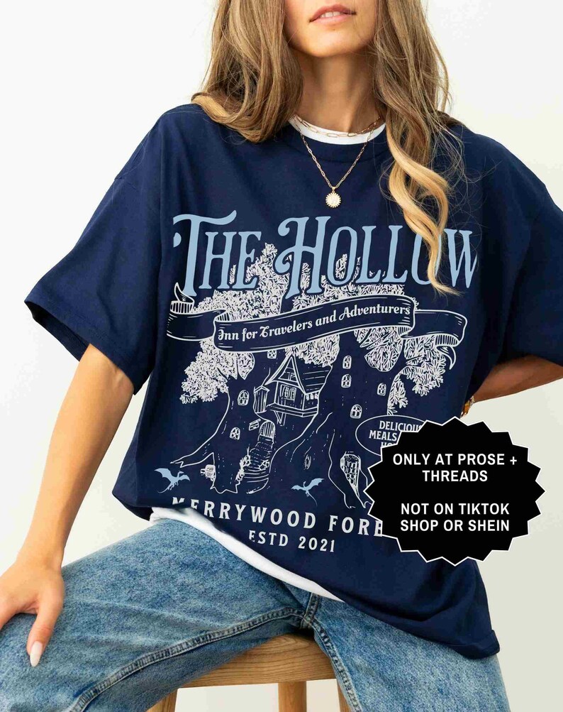 Pu&ograve; includere: T-shirt blu navy con colletto girocollo bianco. La maglietta presenta una grafica azzurra con la scritta "THE HOLLOW Inn for Travelers and Adventurers" e "MERRYWOOD FOR ESTD 2021". Un adesivo in bianco e nero &egrave; sul lato destro.