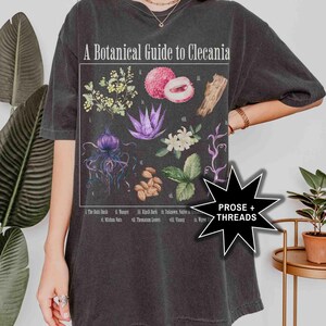 May include: A black t-shirt with a botanical illustration of plants and flowers. The illustration is titled "A Botanical Guide to Clecania" and includes the names of each plant. The t-shirt also has the text "PROSE + THREADS" on the bottom right corner.