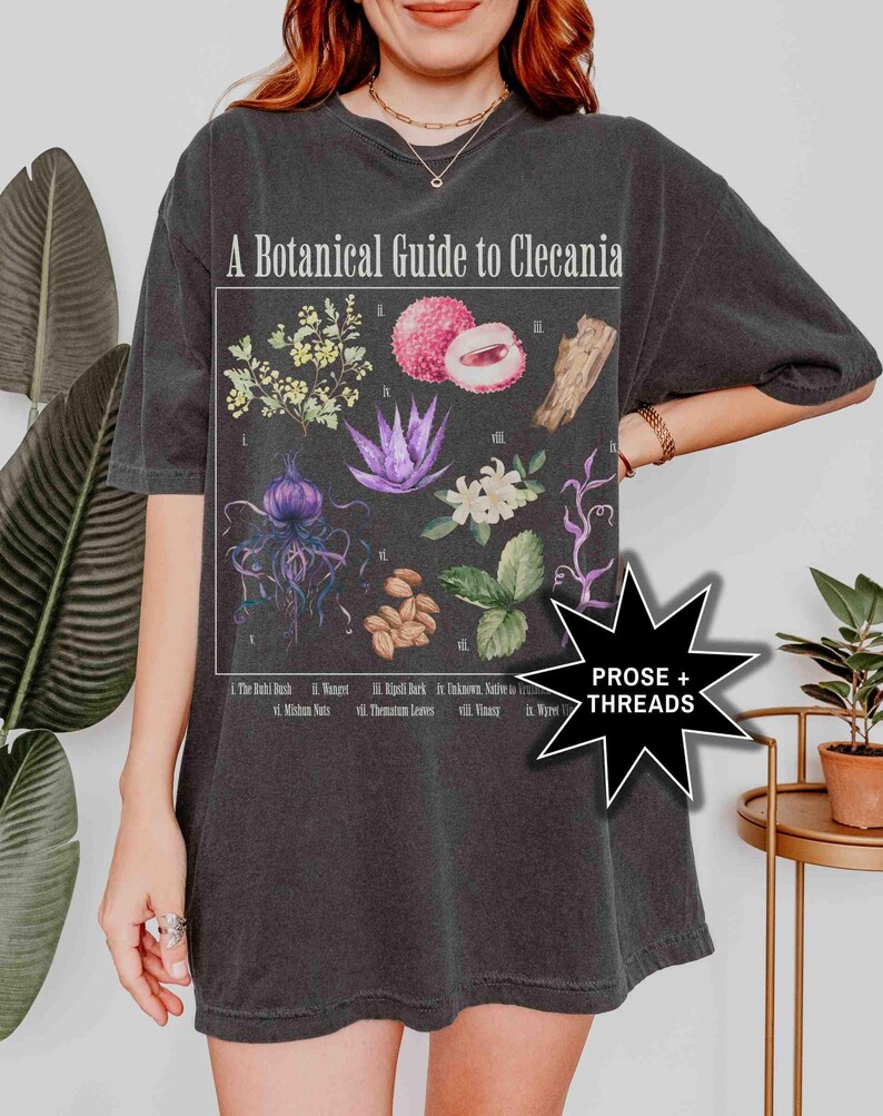 May include: A black t-shirt with a botanical illustration of plants and flowers. The illustration is titled "A Botanical Guide to Clecania" and includes the names of each plant. The t-shirt also has the text "PROSE + THREADS" on the bottom right corner.