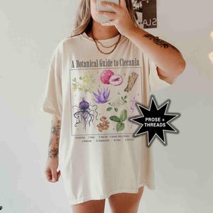 May include: A white t-shirt with a botanical illustration and the text "A Botanical Guide to Clecania". The illustration features a variety of plants and flowers, including a purple flower, a red fruit, and a brown seed pod. The text "PROSE + THREADS" is visible in a black star shape.