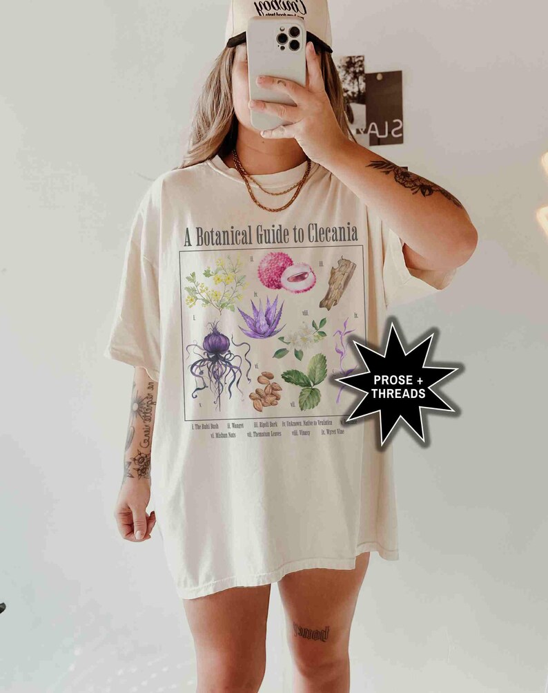 May include: A white t-shirt with a botanical illustration and the text "A Botanical Guide to Clecania". The illustration features a variety of plants and flowers, including a purple flower, a red fruit, and a brown seed pod. The text "PROSE + THREADS" is visible in a black star shape.