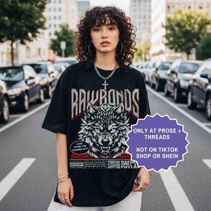 Op de afbeelding: Zwart grafisch t-shirt met een wolfontwerp en de tekst "RAWBONDS". Het shirt heeft ook extra tekst, waaronder "LIVE! FROM THE BONDED CITY" en "STRATEGOS DAEMO KRYPTOS PHYLA". Het shirt wordt door een persoon gedragen.