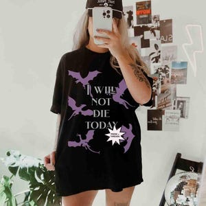 May include: A black t-shirt with a purple graphic of bats and the text "I Will Not Die Today". The graphic also includes a small white star with the text "Prose + Threads" inside.