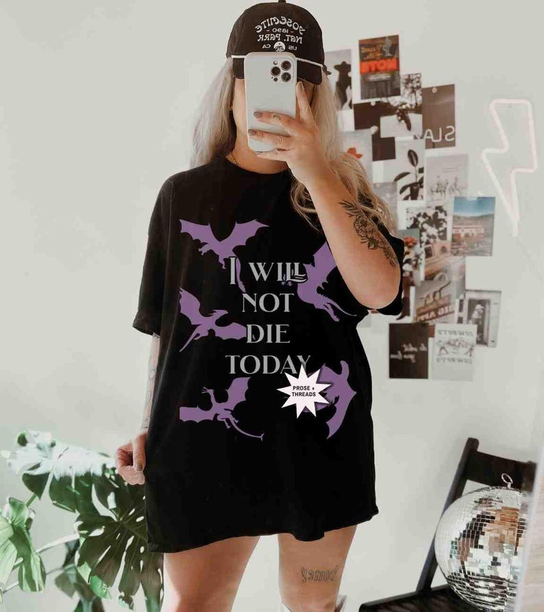 May include: A black t-shirt with a purple graphic of bats and the text "I Will Not Die Today". The graphic also includes a small white star with the text "Prose + Threads" inside.