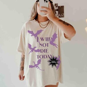 May include: A white t-shirt with a purple dragon graphic and the text "I Will Not Die Today". The shirt also features a black starburst with the text "Prose + Threads".