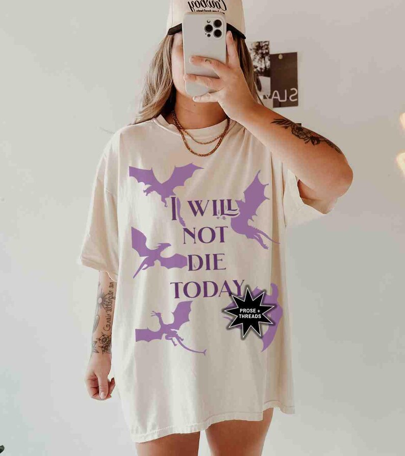 May include: A white t-shirt with a purple dragon graphic and the text "I Will Not Die Today". The shirt also features a black starburst with the text "Prose + Threads".